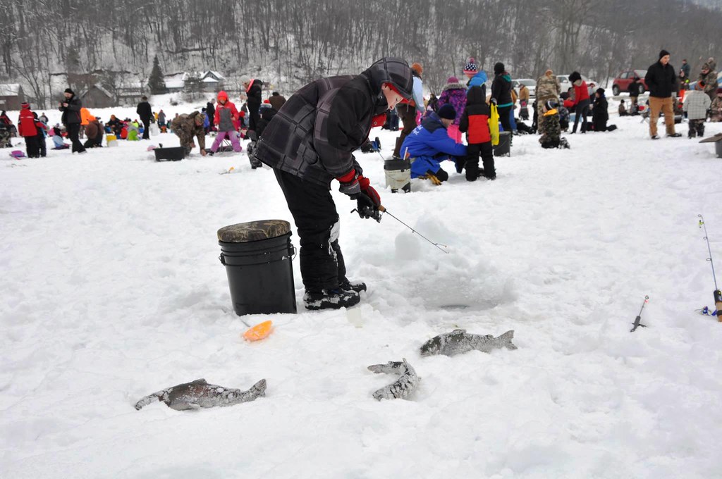 a child dressed in winter gear stands with a fishing pole over a hole in a frozen lake