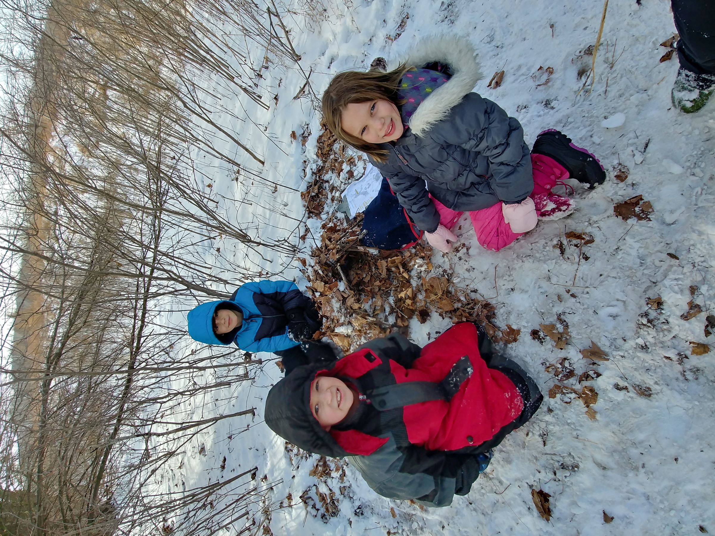 3 kids smile at the camera while sitting around their built animal shelter in winter