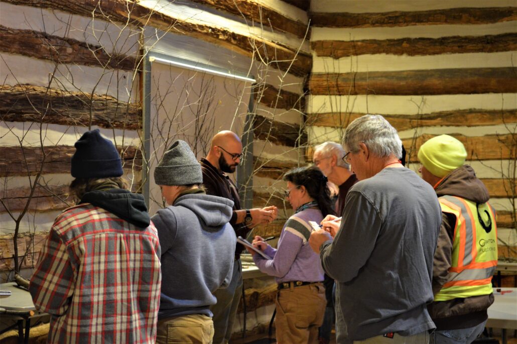 6 people stand in the Riveredge barn taking notes while listening to a presenter and looking at tree branches