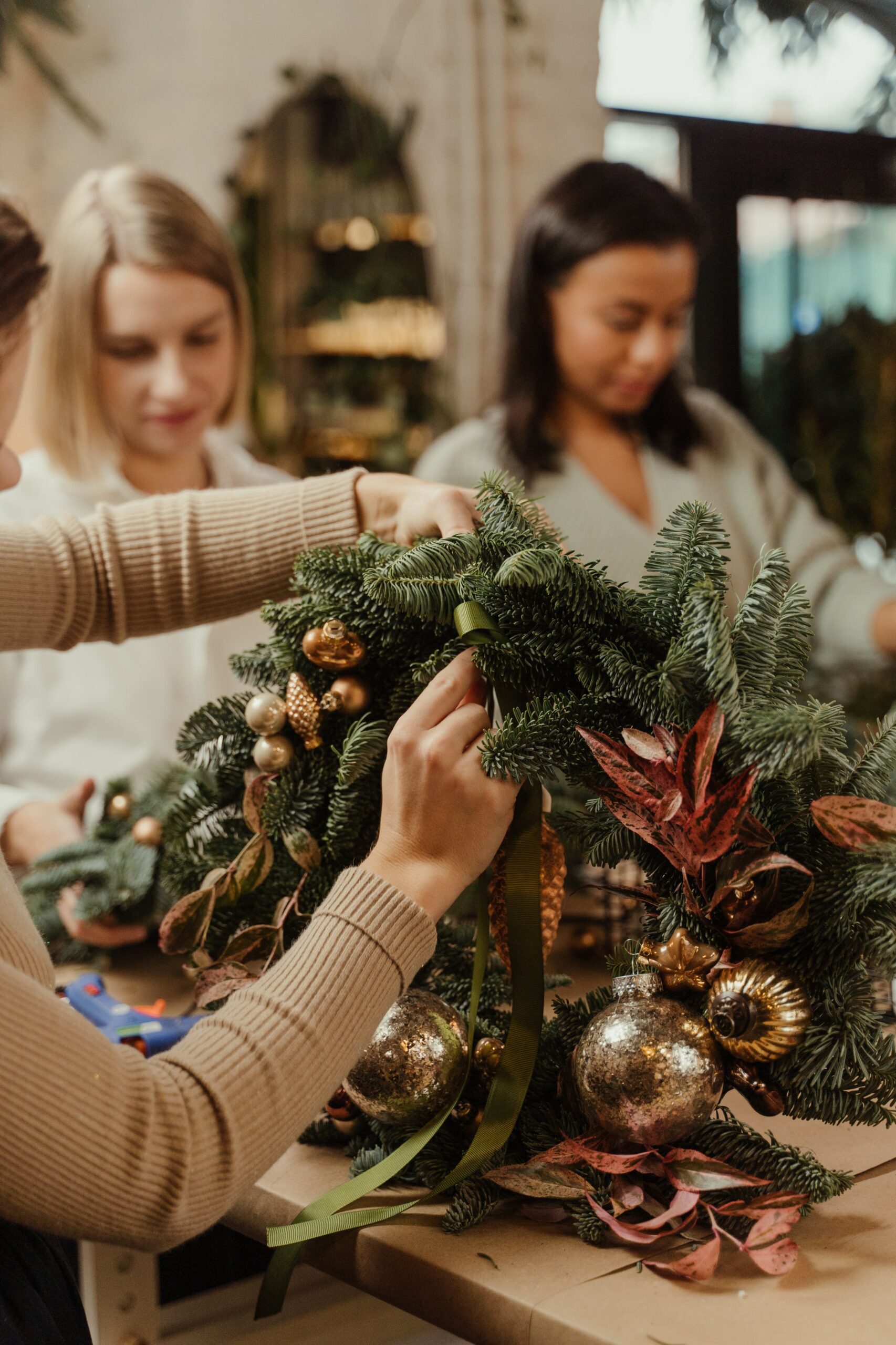 close up of a holiday wreath being made with 2 other people in the background