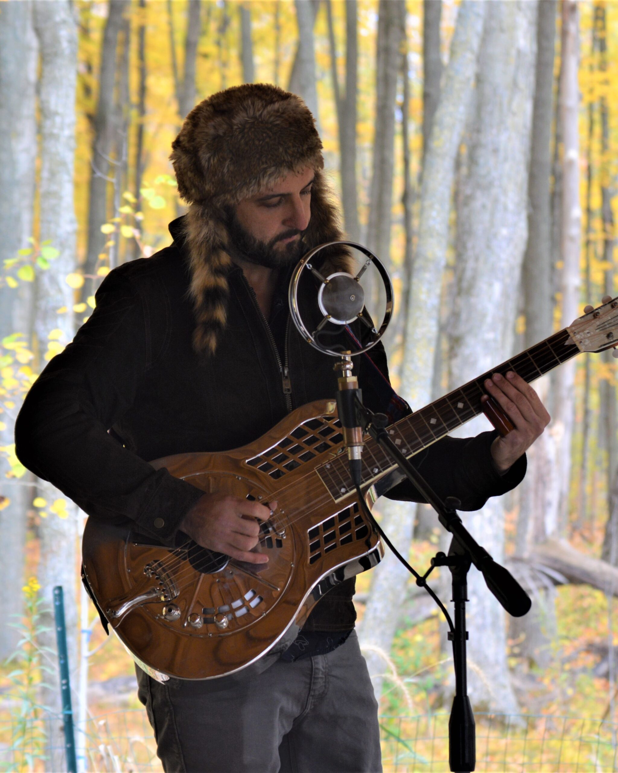 Luke Cerny, a young white man, stands in the autumn woods at Riveredge playing his guitar while singing into a microphone
