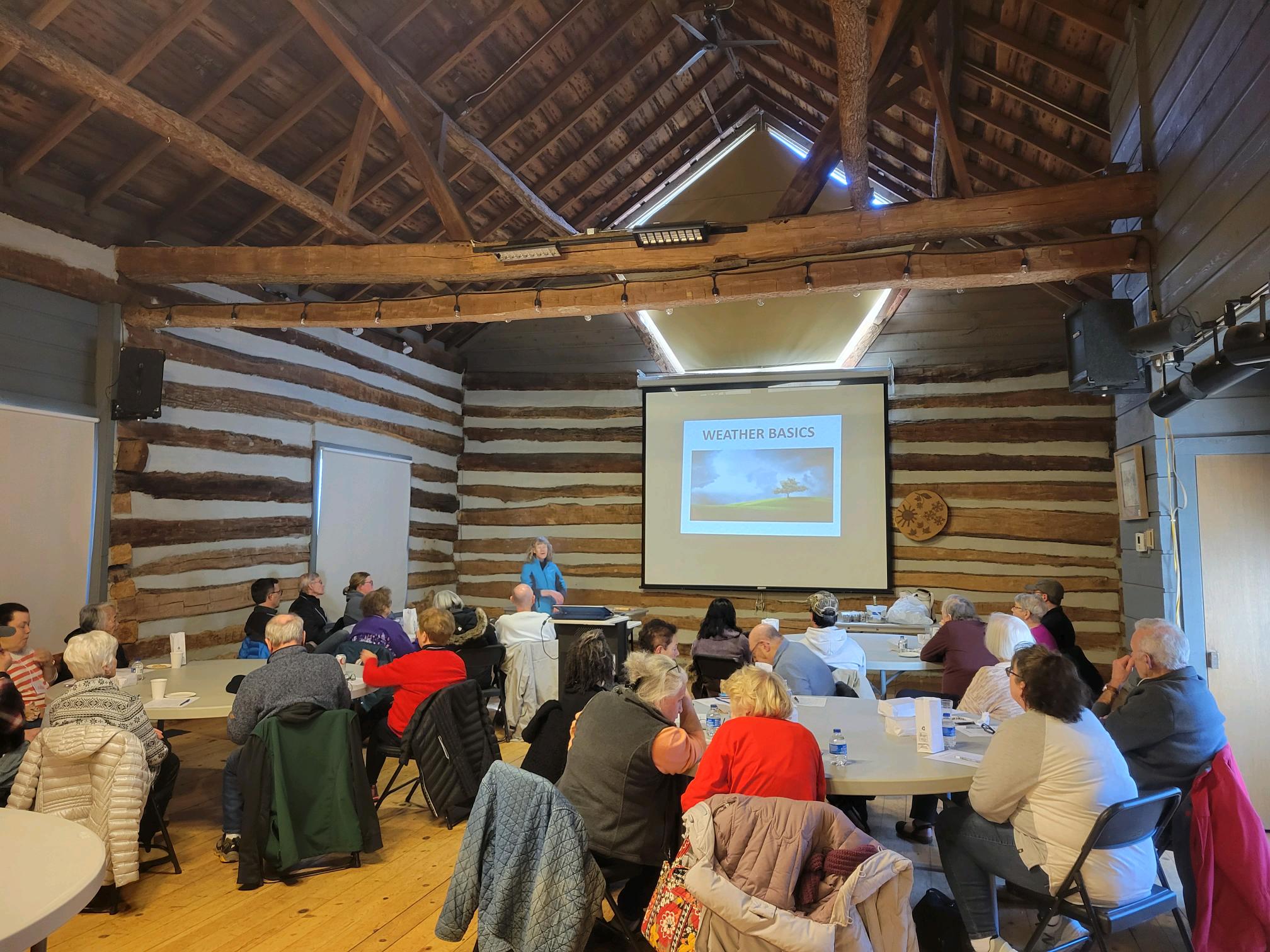 a group of older adults sits at tables in the Riverdge Barns listening to a presentation about weather