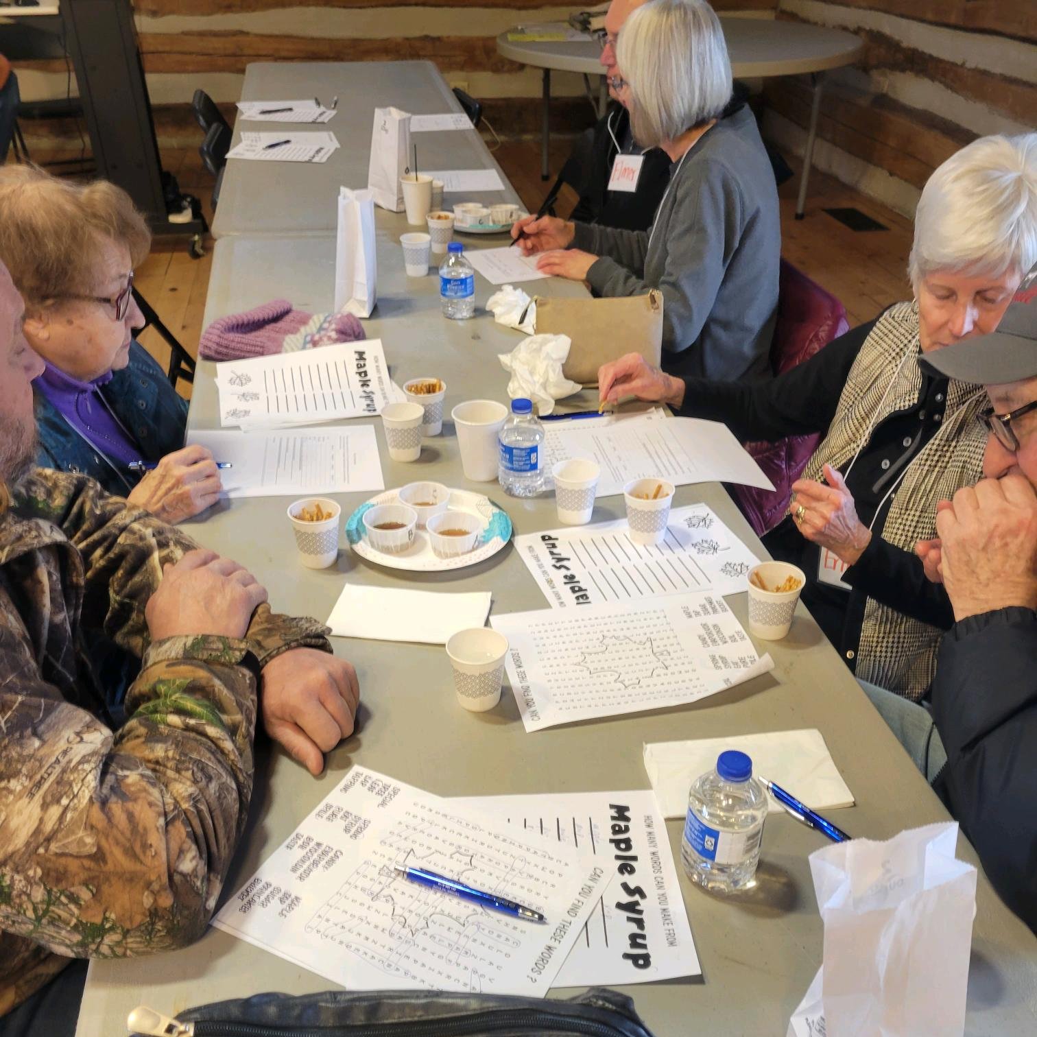 6 older adults sit at a table in the Riveredge barn sampling different types of maple syrup