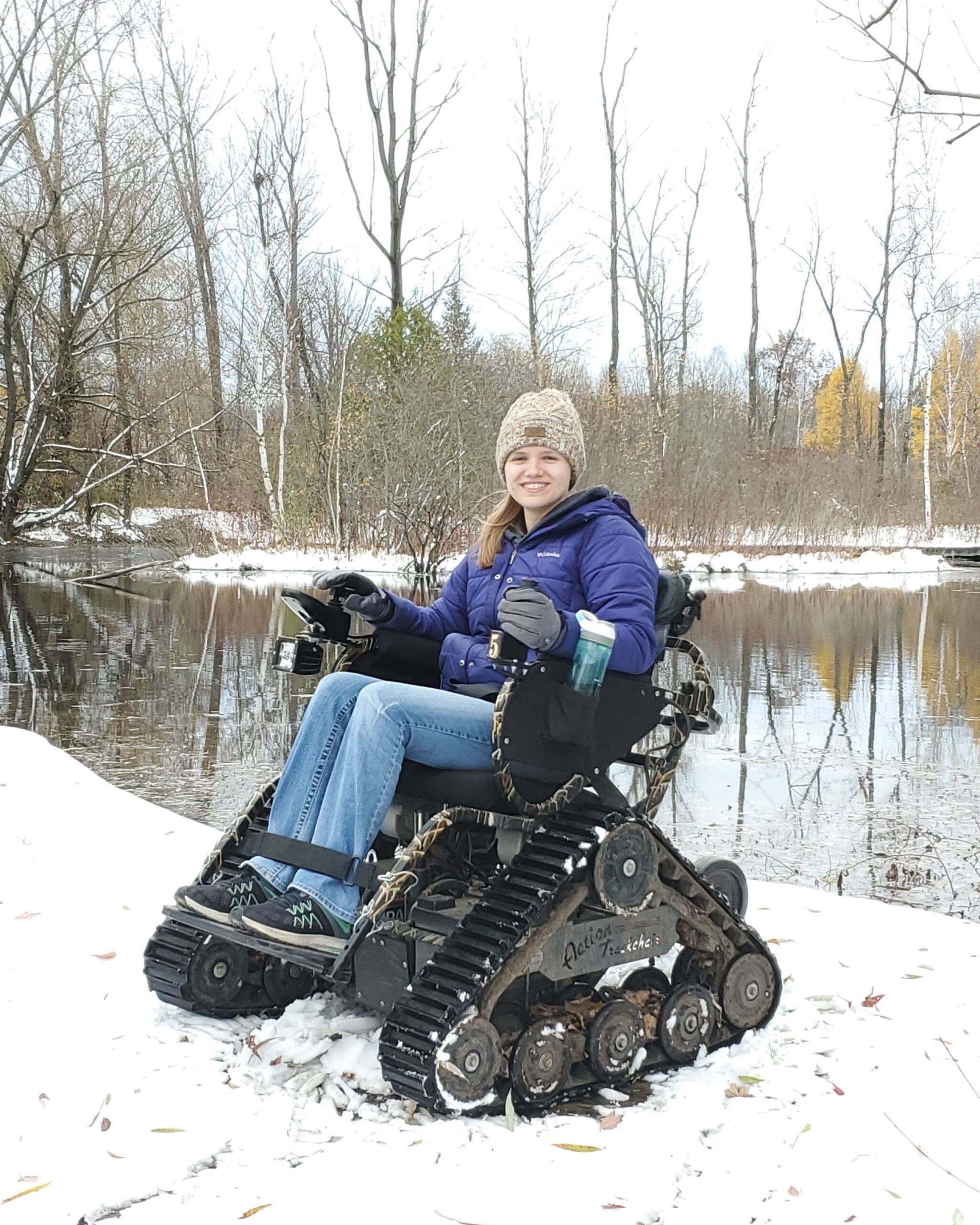 a young woman sits in an all terrain wheelchair in front of a Riveredge pond in winter with snow on the ground