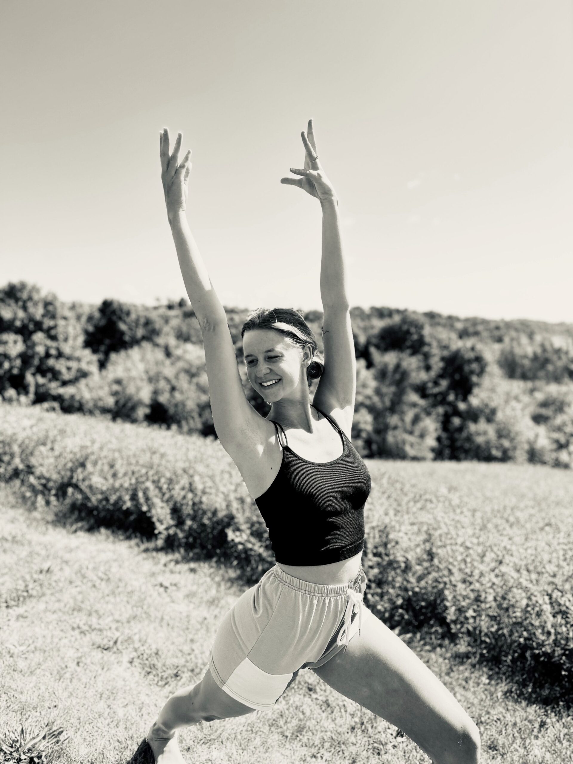black and white photo of Mary Addy, a young white yoga instructor, smiling and doing a yoga pose reaching toward the sky with grass and trees in the background