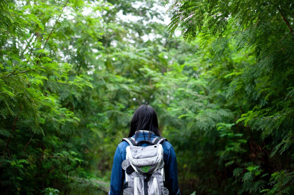 a person with long black hair facing away from the camera wearing a backpack in a lush green forest