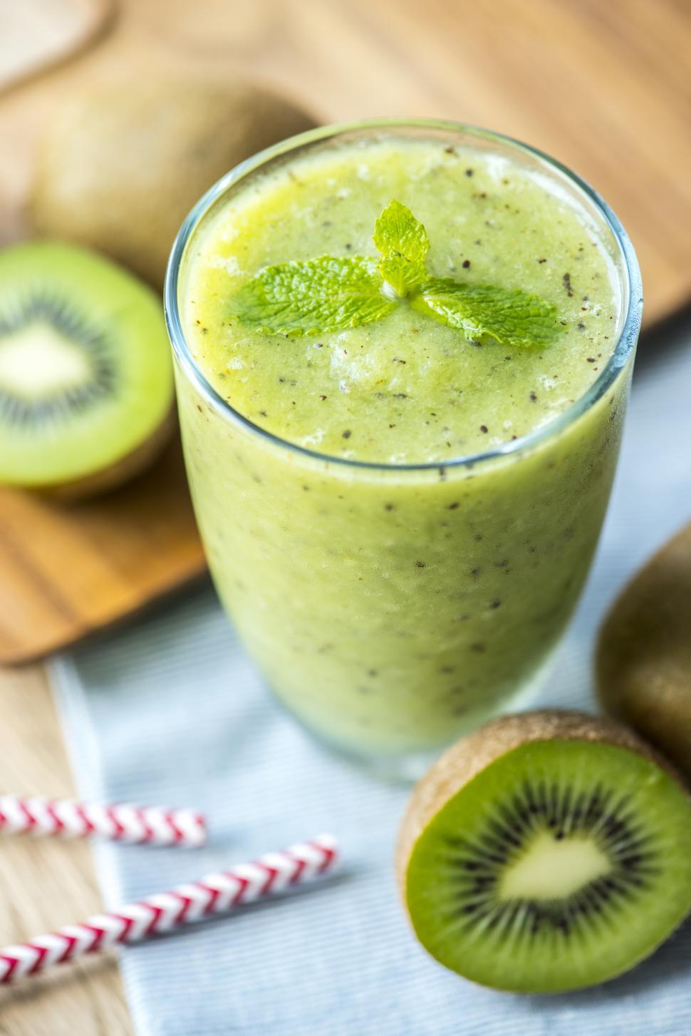 close up of a green smoothie in a glass cup with kiwi and straws on the table next to it