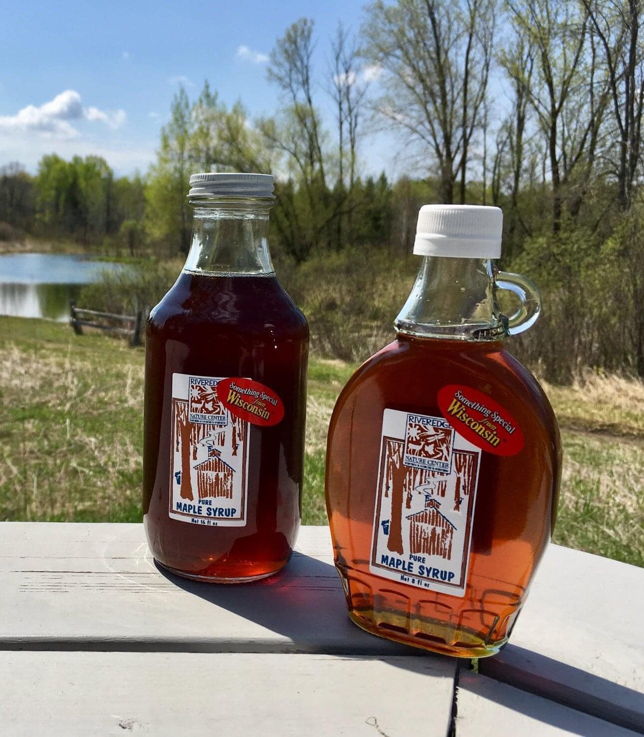 close up of Riveredge Maple Syrup bottles with a springtime pond and trees in the background