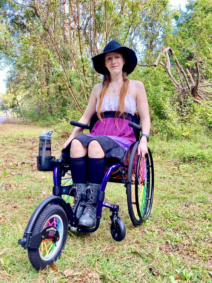 a young adult in a wheelchair with rainbow spokes and a large front wheel attachment on a trail with trees in the backgorund