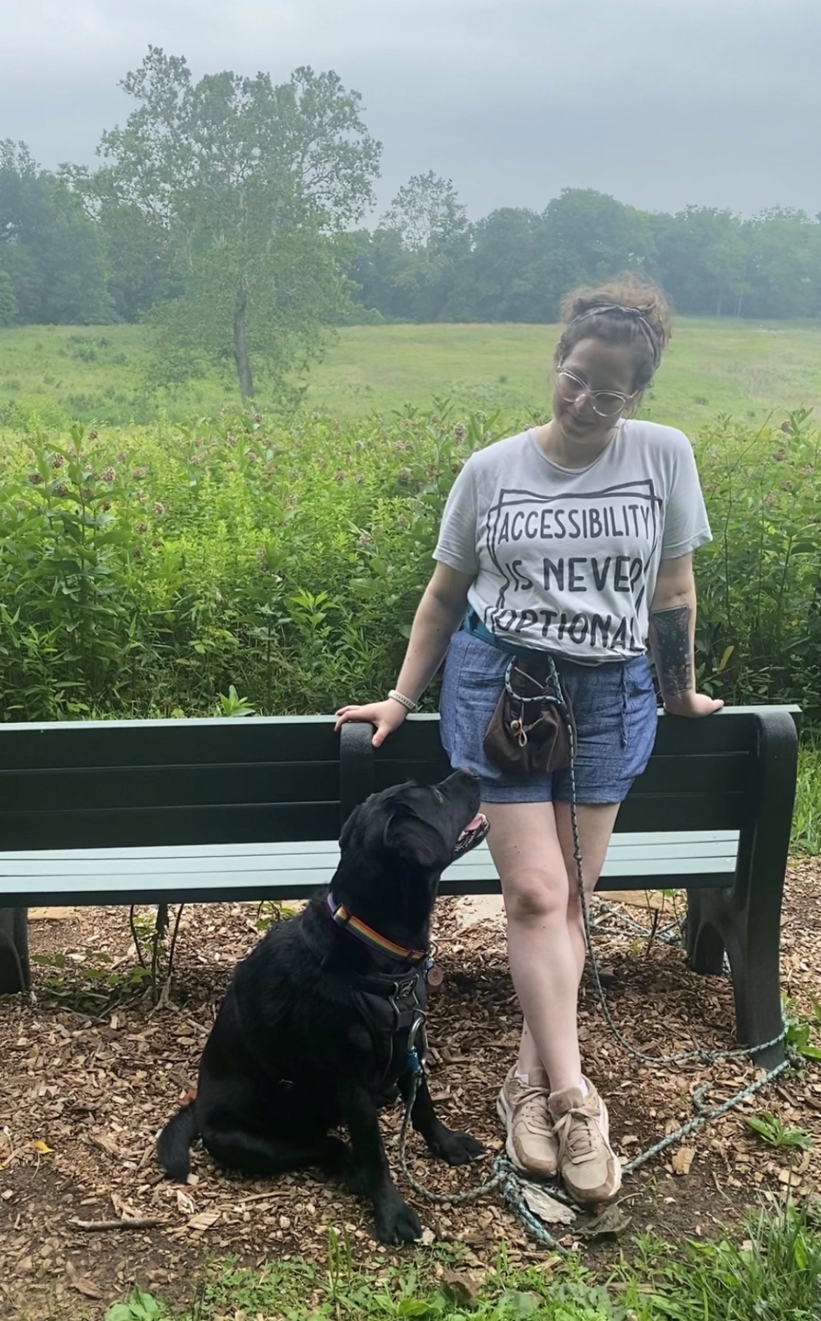 a white woman standing outside leaning on a bench and looking down at her large black service dog who is looking up at her